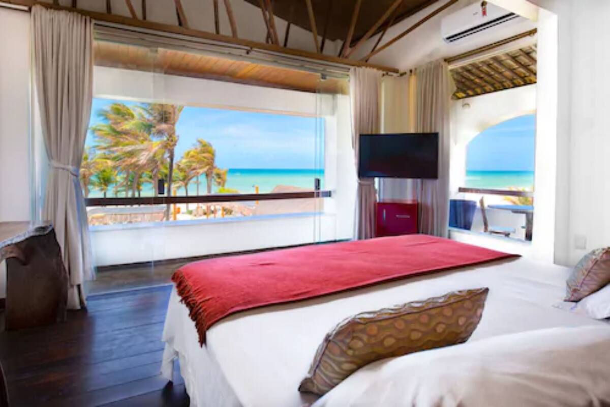 A hotel room with a large bed, a TV, and a balcony with a view of the beach and palm trees.