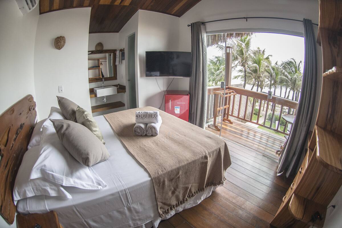 A bedroom with a bed, pillows, a TV, a red cabinet, and a balcony with a view of palm trees.