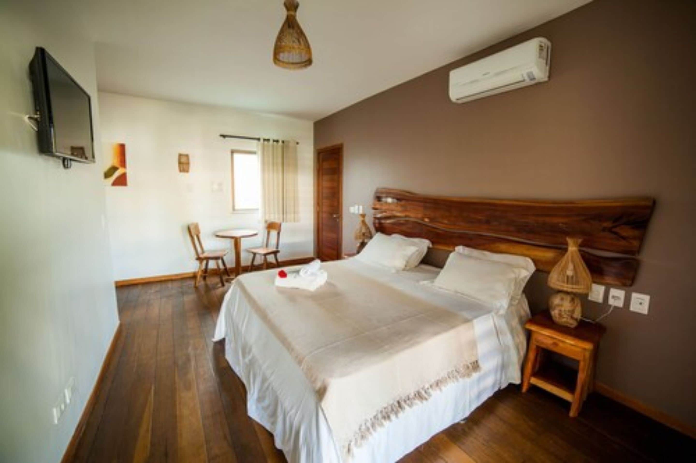 A bedroom with a large bed, a wooden headboard, a bedside table with a lamp, a TV mounted on the wall, and a small table with chairs in the corner.