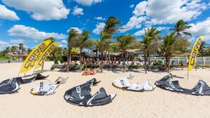 A beach with multiple windsurfing boards and flags, palm trees, and people relaxing on the sand. - Vila Coqueiros (Caucaia)