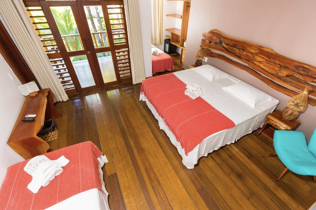 A hotel room with two beds, wooden flooring, a wooden headboard, a small table, a chair, and a balcony with a view of greenery.