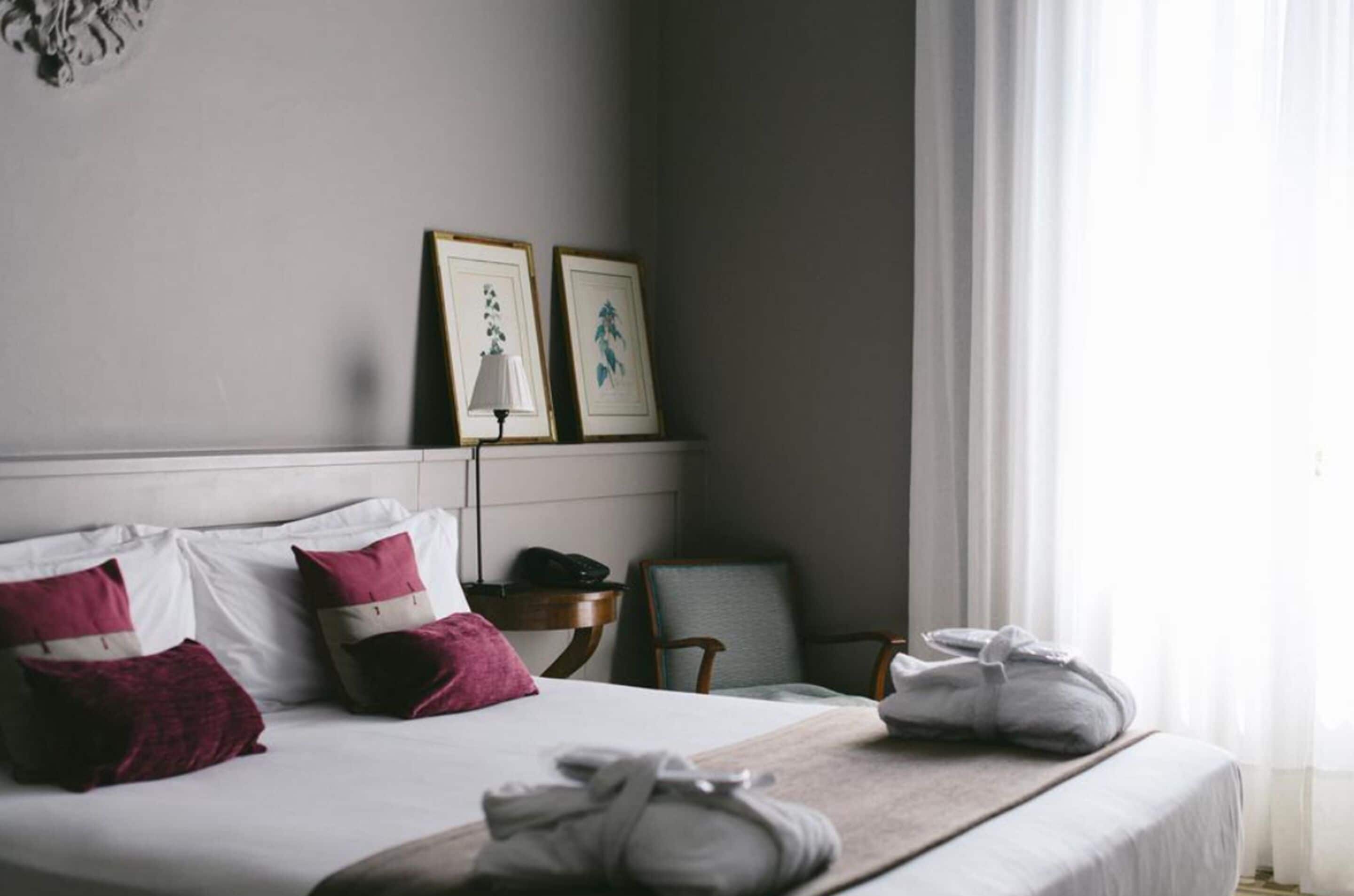 Una cama bien tendida con almohadas blancas y rosas, una mesita de noche con una lámpara y una silla.