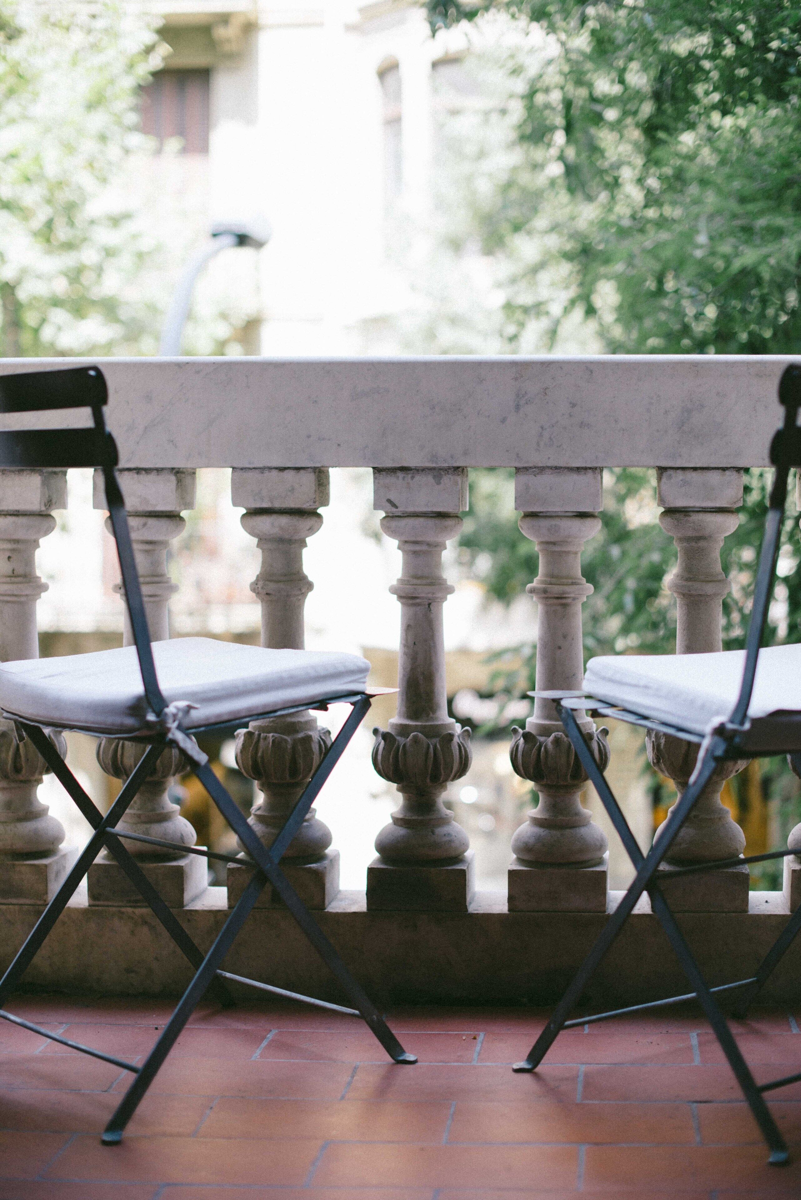 A balcony with two chairs and a table, featuring a stone balustrade and a view of greenery.