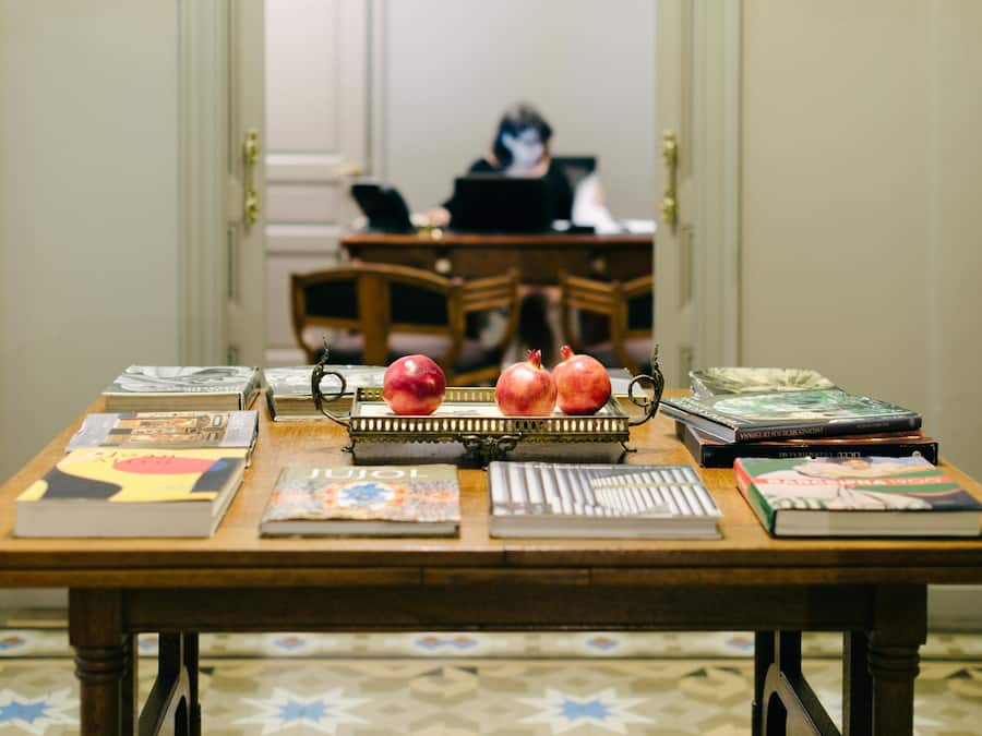 A wooden table with books, a tray of pomegranates, and a laptop in a room with patterned floor tiles.
