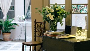 Una habitación con piso estampado, una silla de madera, una maleta negra, un florero con flores y una mesa con libros y una lámpara.