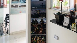 A vertical display of posters with various beach scenes and activities, including 'PRAIA DO JABAQUARA', 'NASCER DO SOL', 'ESCUNA', 'CAIAQUE', 'STAND UP', 'BLOCO DA LAMA', and 'ABRASTÃO DO JABAQUARA'. - Pousada Mar Azul (Paraty)