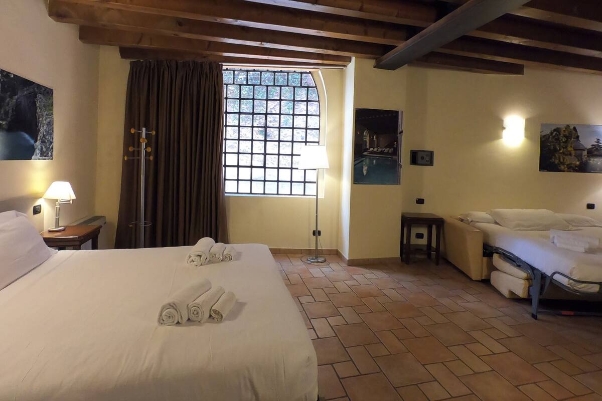 Habitación de hotel con una cama grande, una cama más pequeña, una ventana con cortinas, una lámpara y obras de arte en las paredes.