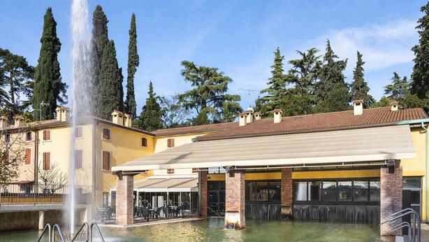 Un espace piscine agrémenté d’une fontaine, entouré de bâtiments dotés de terrasses extérieures et d’un patio couvert.