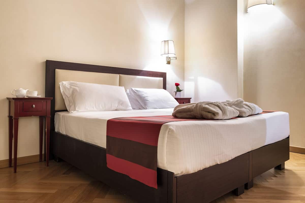 Una cama bien hecha con sábanas blancas y una manta a rayas marrones y rojas, una mesita de noche de madera con una taza y un platillo, y una lámpara fijada a la pared.