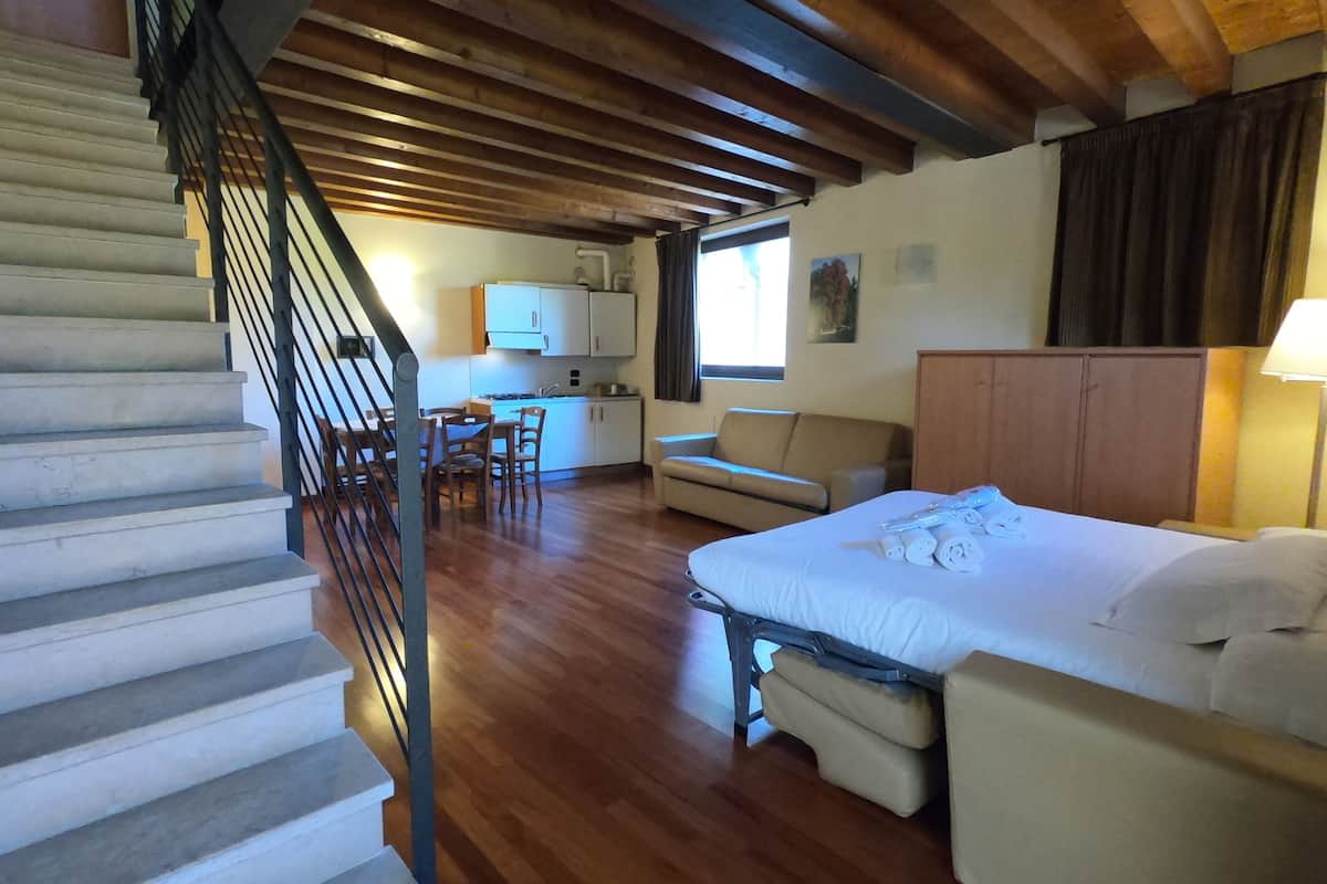 Habitación de hotel con escalera de madera, cama con toallas enrolladas, sofá, pequeña cocina y mesa de comedor.