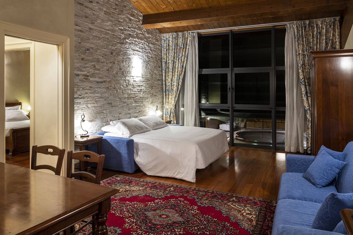 Un dormitorio con pared de piedra, una cama, un sofá azul, una mesa de madera y vistas a un jacuzzi exterior.
