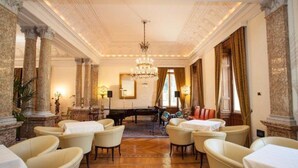 Une vaste pièce ornée de colonnes de marbre, d’un lustre et d’un piano.