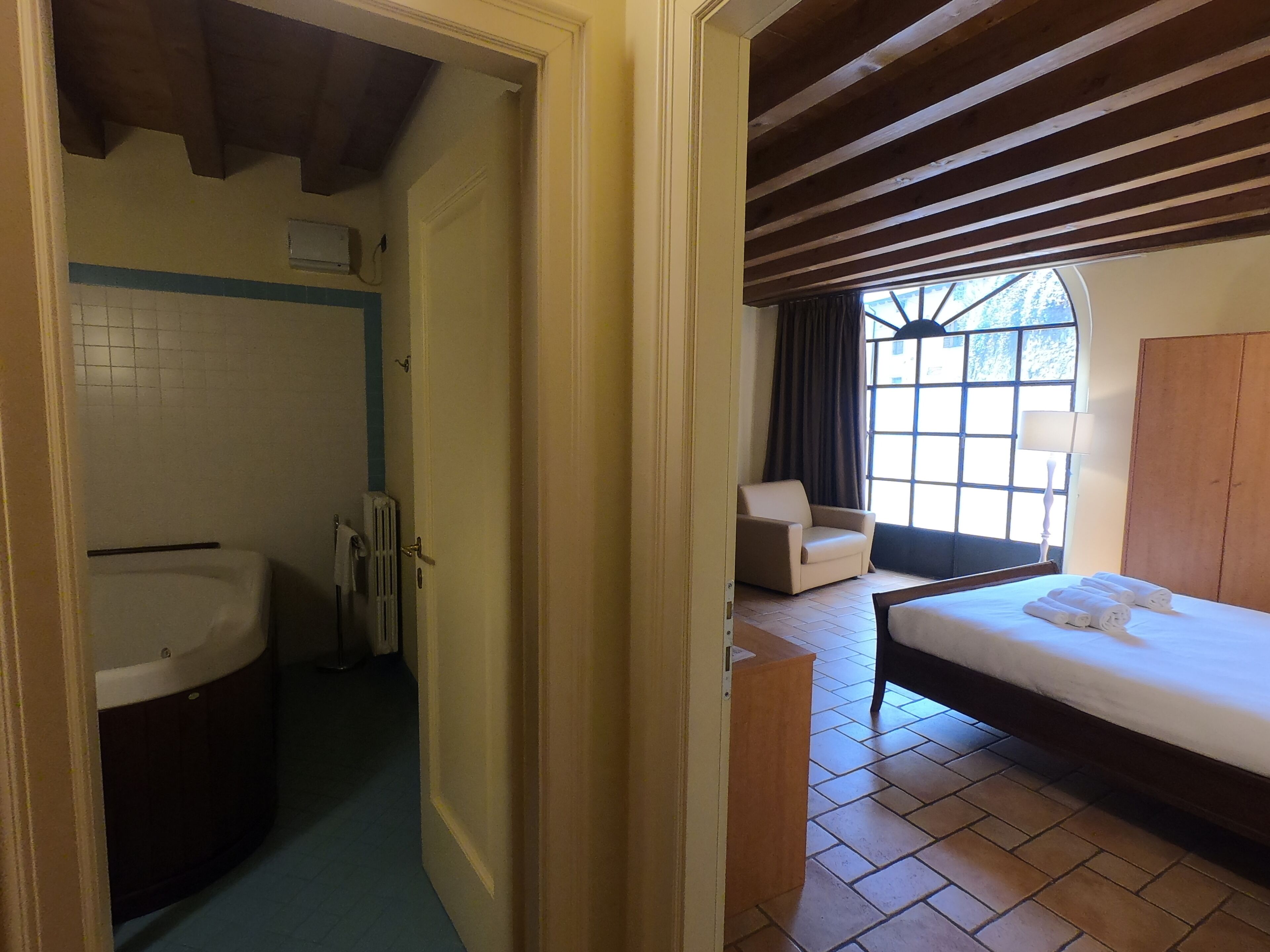 A bedroom with a bed, a chair, a window, and a bathroom with a bathtub and radiator.