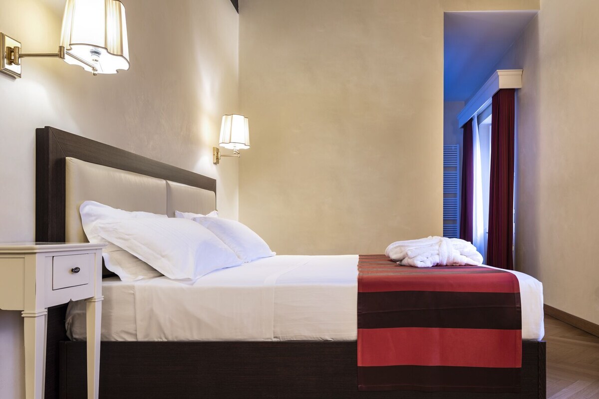 A neatly made bed with white linens, a dark wooden headboard, and a red and black throw blanket. There is a white bedside table with a drawer, and two wall-mounted lamps provide lighting.