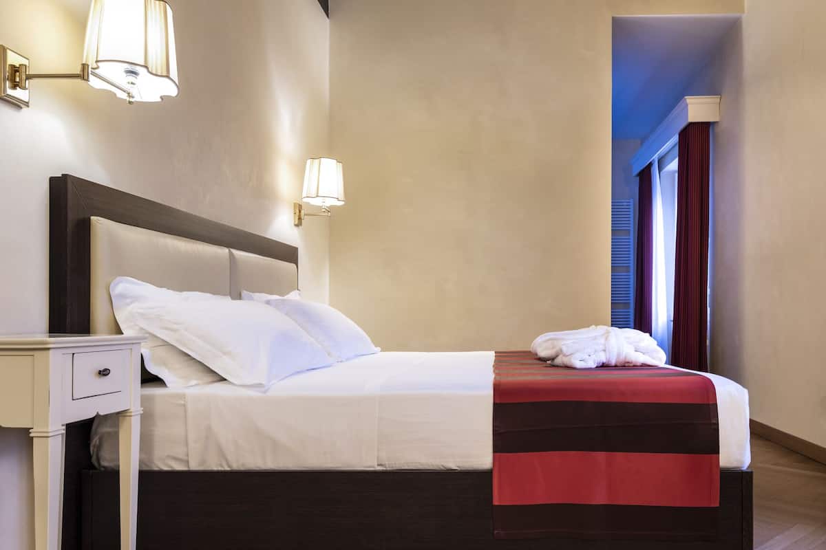 Una cama bien hecha con sábanas blancas, cabecera de madera oscura y una manta de color rojo y negro. Hay una mesita de noche blanca con cajón y una lámpara de pared. La habitación tiene suelo de madera y una ventana con cortinas rojas.