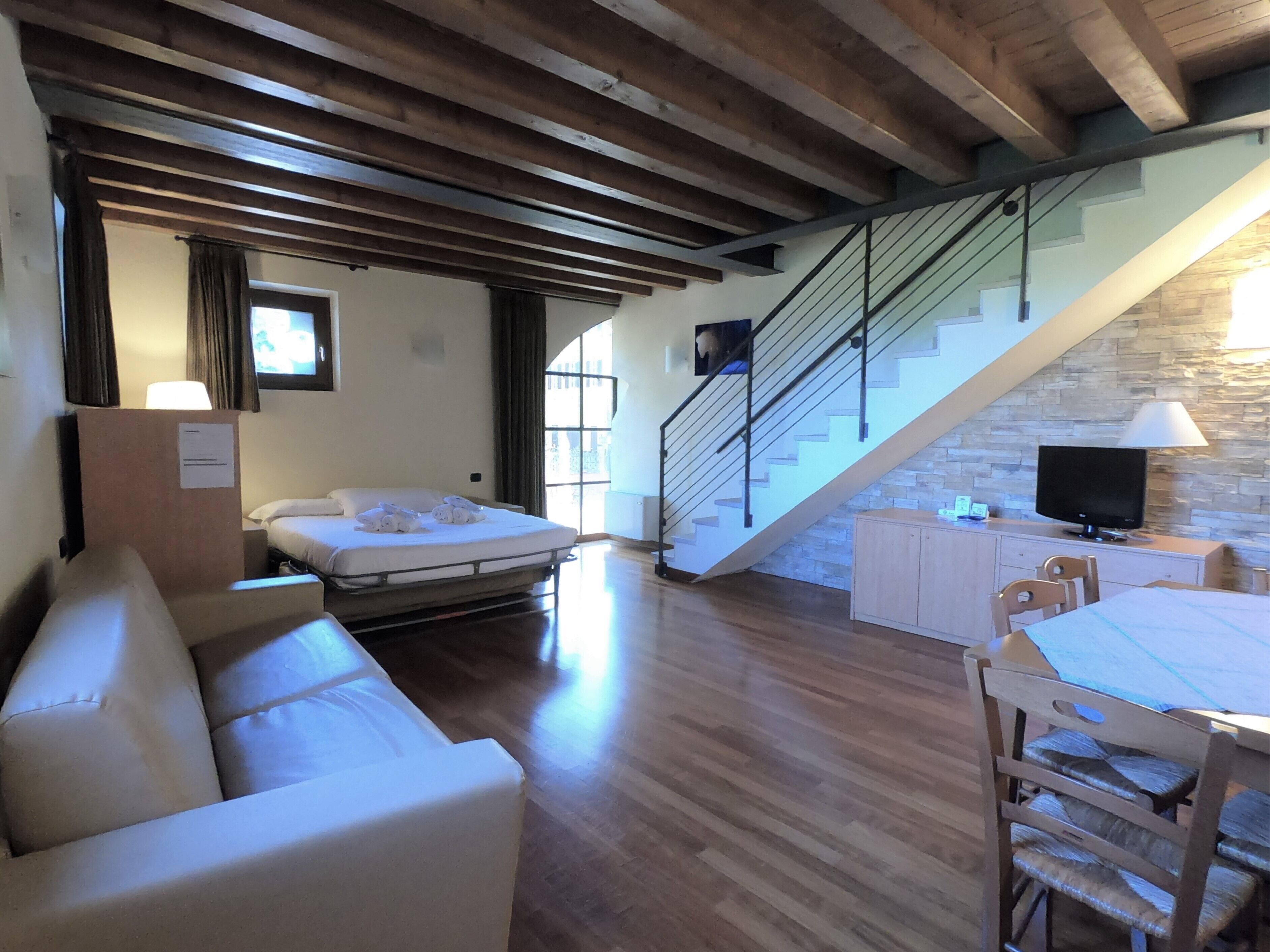 Habitación de hotel con techo de madera, un sofá, una cama, una escalera, un televisor y una pared de piedra.