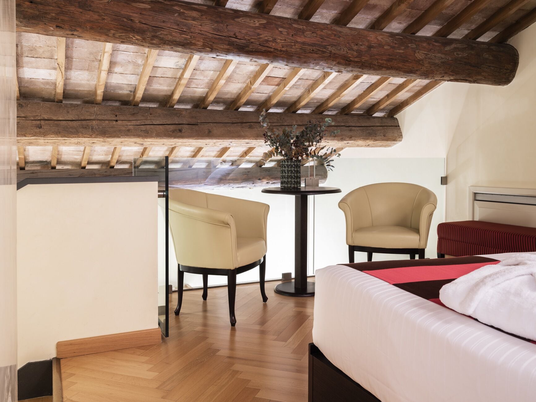 A bedroom with a wooden ceiling, a bed, two chairs, and a small table with a vase.