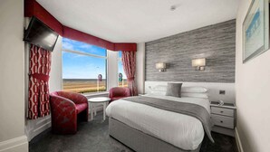 A hotel room with a large bed, two bedside tables, a desk, and a chair. There are two windows with curtains, a view of the beach, and a wall-mounted TV.