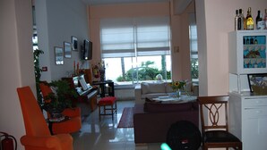 Una sala de estar con sofá, sillas, una mesa de centro y televisión. Hay una ventana con persianas, una estantería y una planta.