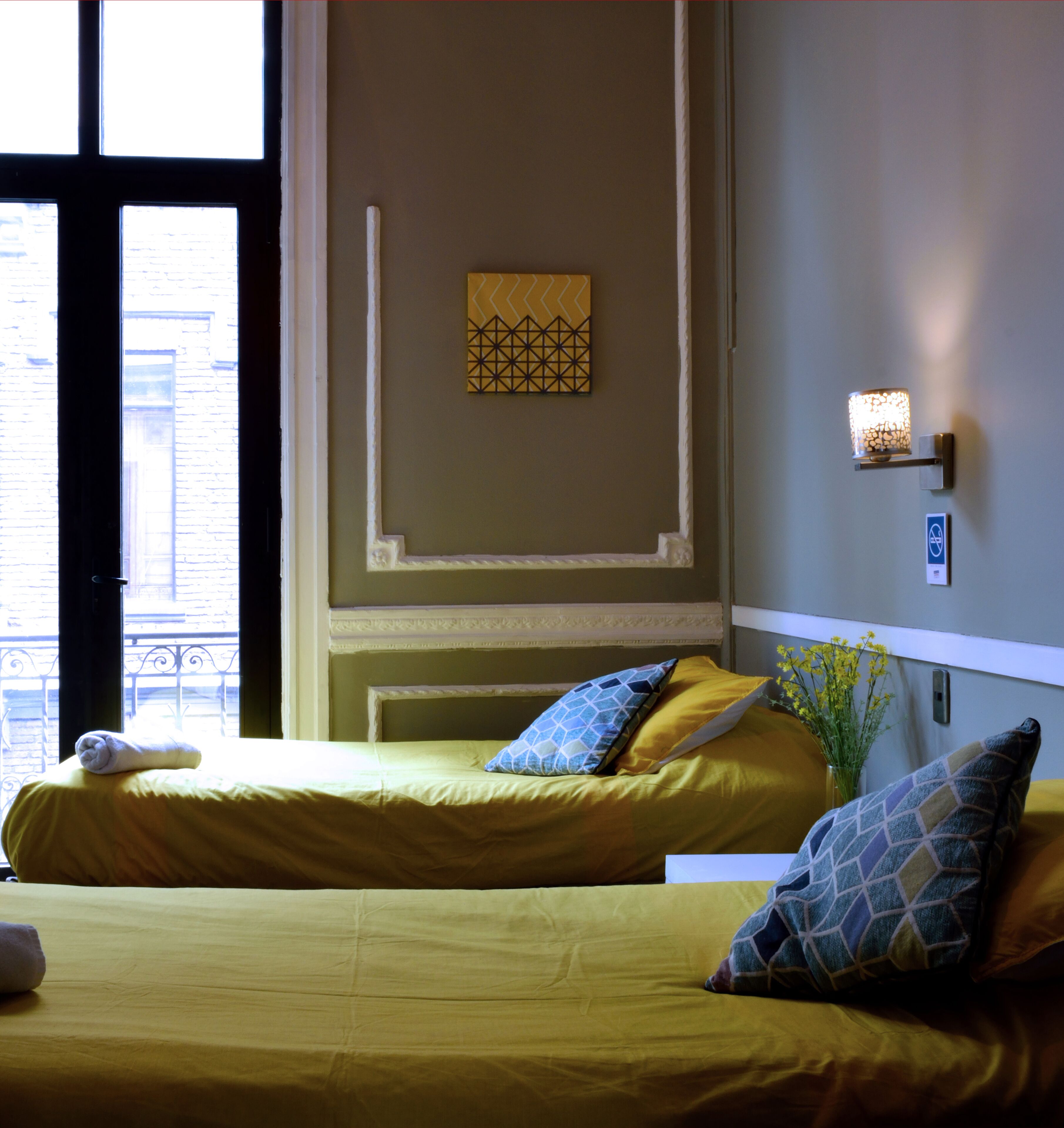 A hotel room with two beds, a decorative wall piece, a wall-mounted light, and a view of the city through large windows.