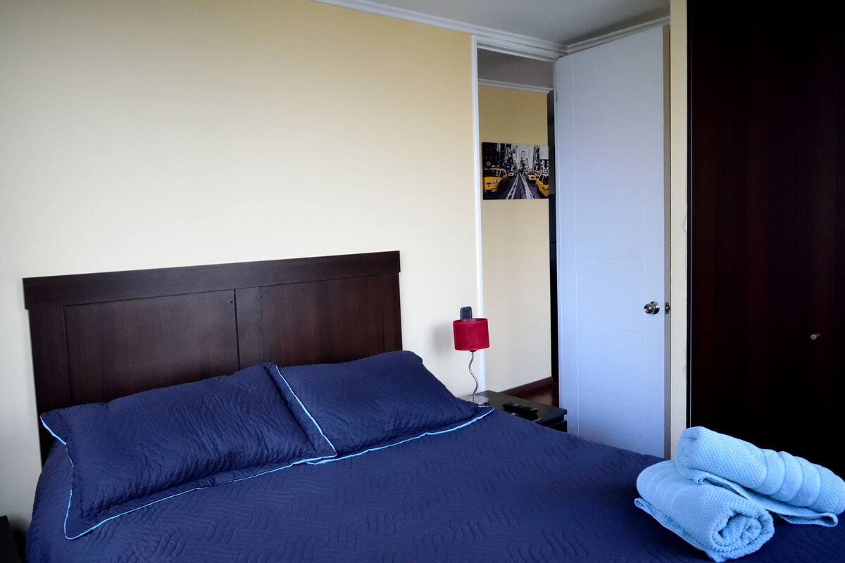 Una cama bien hecha con una colcha azul oscuro y almohadas, una lámpara roja sobre una mesita de noche y una puerta a la derecha.