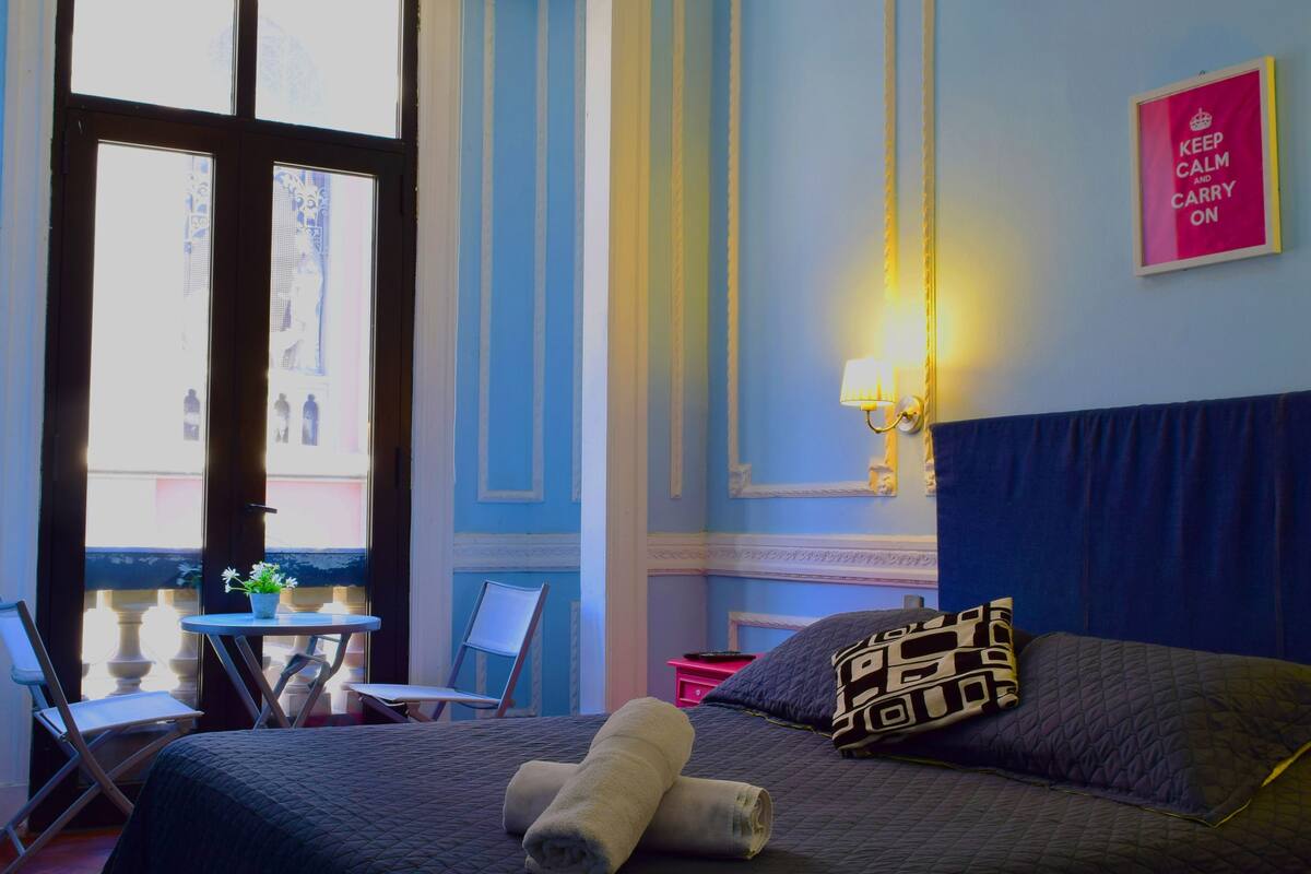 Habitación de hotel con cama, una pequeña mesa con silla, ventana con vistas y una lámpara de pared.
