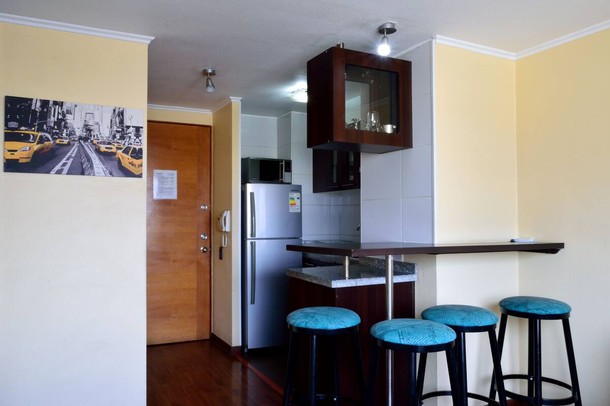 Cocina compacta con refrigerador, microondas y un mueble de madera sobre la encimera. Hay tres taburetes azules y un espejo montado en la pared.