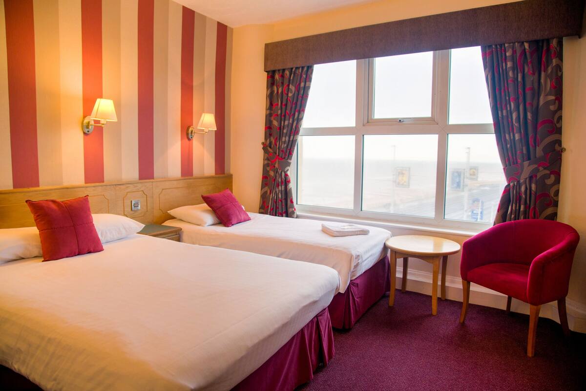 Une chambre d’hôtel avec deux lits, une petite table, une chaise rouge et une fenêtre avec des rideaux.