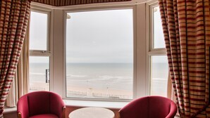 Ein Zimmer mit Erkerfenster mit Blick auf einen Strand, ausgestattet mit zwei roten Sesseln und einem kleinen runden Tisch.