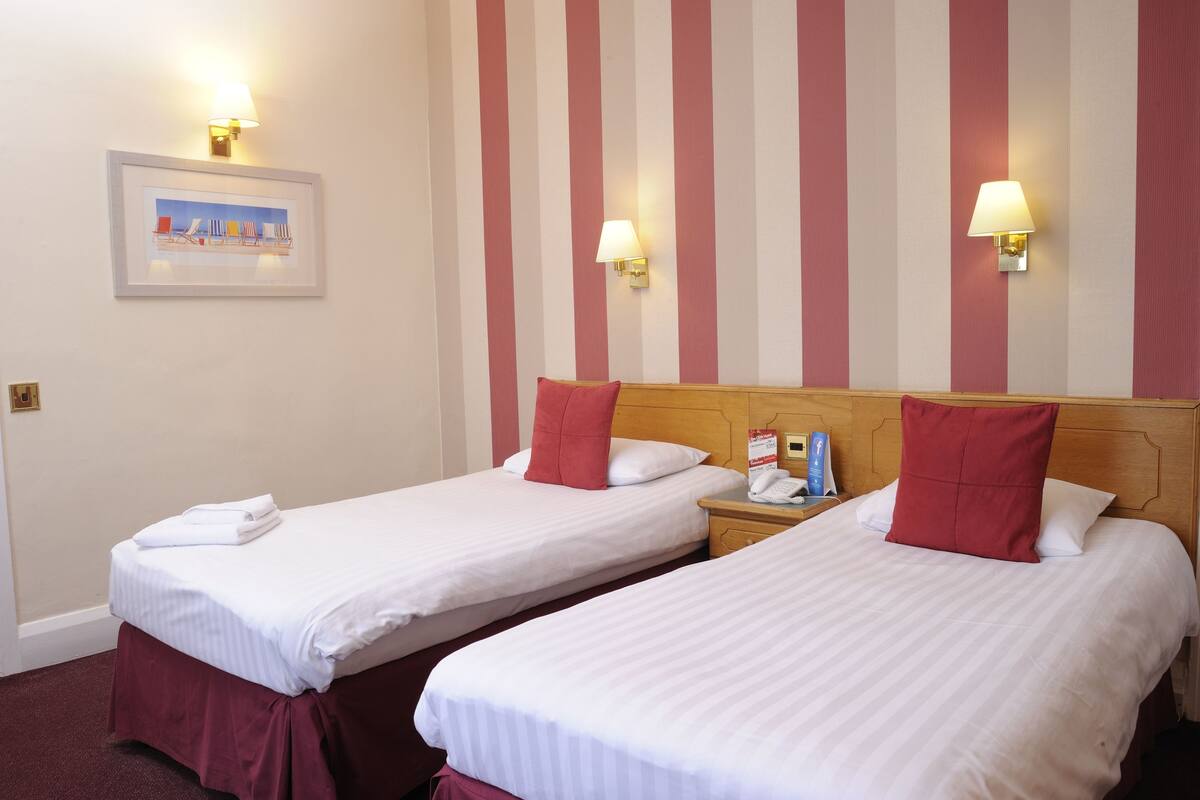 Deux lits simples avec du linge de lit blanc et des oreillers rouges, un tableau encadré au mur et deux lampes fixées au mur.