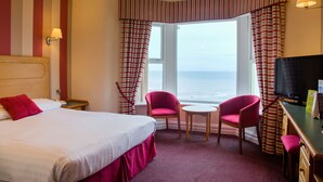 Ein Hotelzimmer mit Bett, zwei Sessel, einem kleinen Tisch, einem Fernseher und einem Fenster mit Vorhängen.