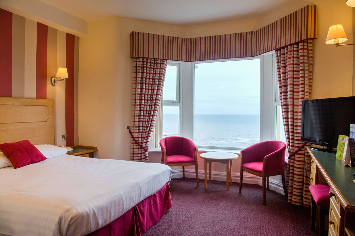 Une chambre d’hôtel avec un lit, deux chaises, une petite table, un téléviseur et une fenêtre avec des rideaux.