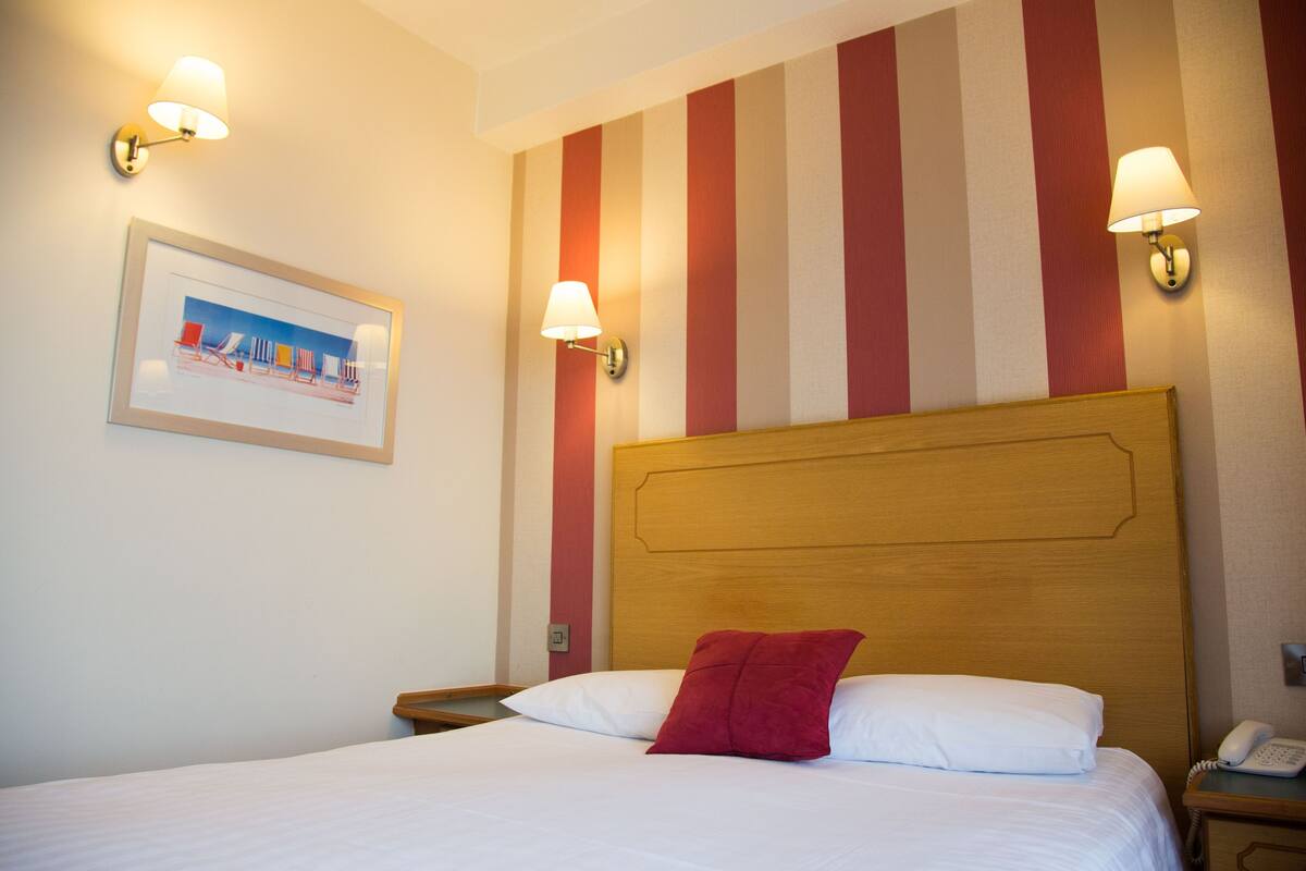 Une chambre d’hôtel avec une tête de lit en bois, un lit avec une literie blanche et un oreiller rouge, deux tables de chevet avec des lampes et un tableau encadré au mur.