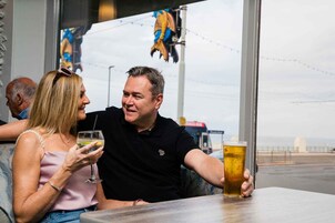 Twee personen zitten aan een tafel met drankjes, met uitzicht op een brug en water.