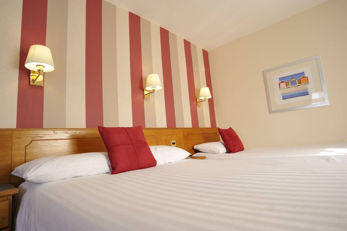 Une chambre d’hôtel avec un grand lit, deux oreillers rouges, une tête de lit en bois et un mur à rayures agrémenté d’appliques murales.