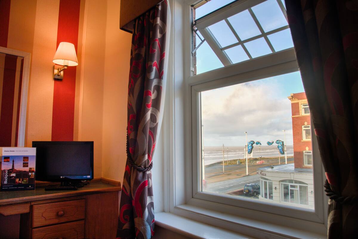Habitación de hotel con una televisión sobre un escritorio de madera, una ventana con vistas a la playa y edificios, y una lámpara fijada a la pared.