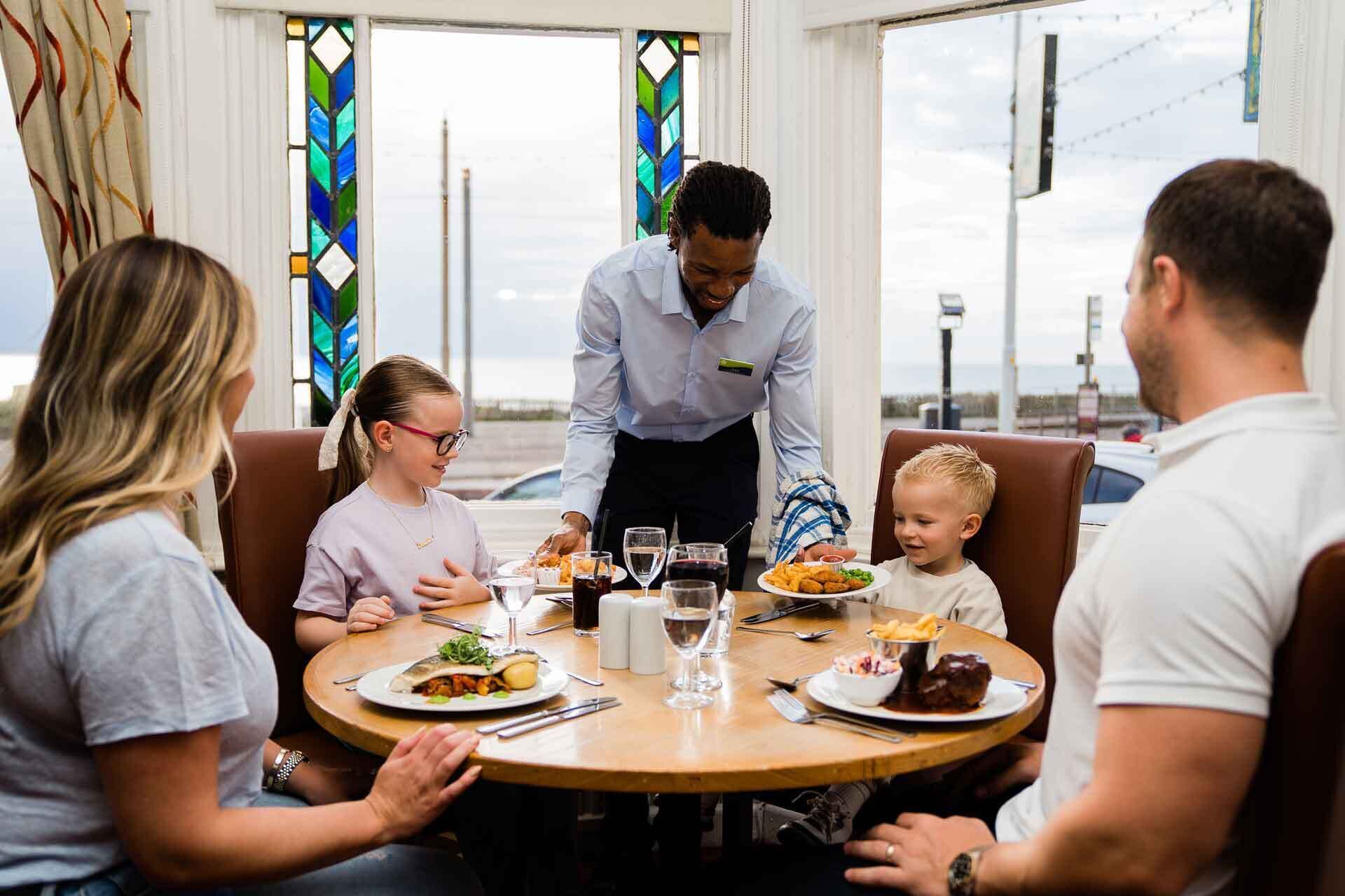 A family dining at a table with a waiter, in a room with large windows and a view of the outside.
