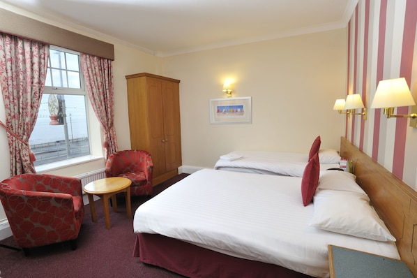 A hotel room with two beds, a wooden wardrobe, a red armchair, a small round table, and a window with curtains. - Cliffs Hotel - Blackpool (Blackpool)