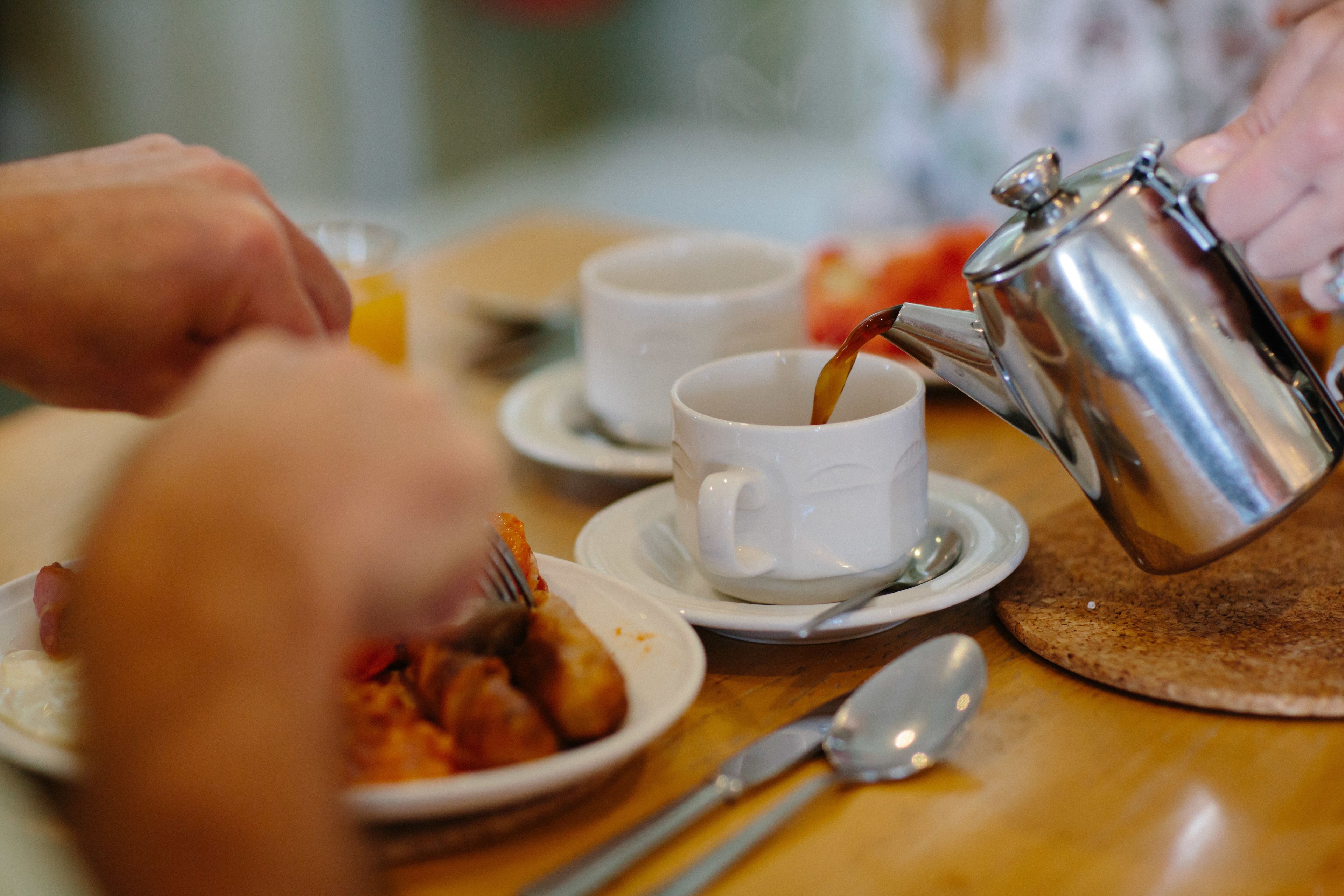 Une table en bois dressée pour le petit-déjeuner, avec une cafetière versant du café dans une tasse, une assiette remplie de nourriture et des couverts.