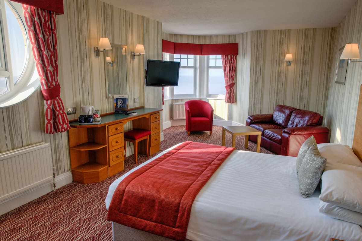 Habitación de hotel con cama, escritorio, sillón rojo, sofá rojo, televisión y ventana con cortinas.