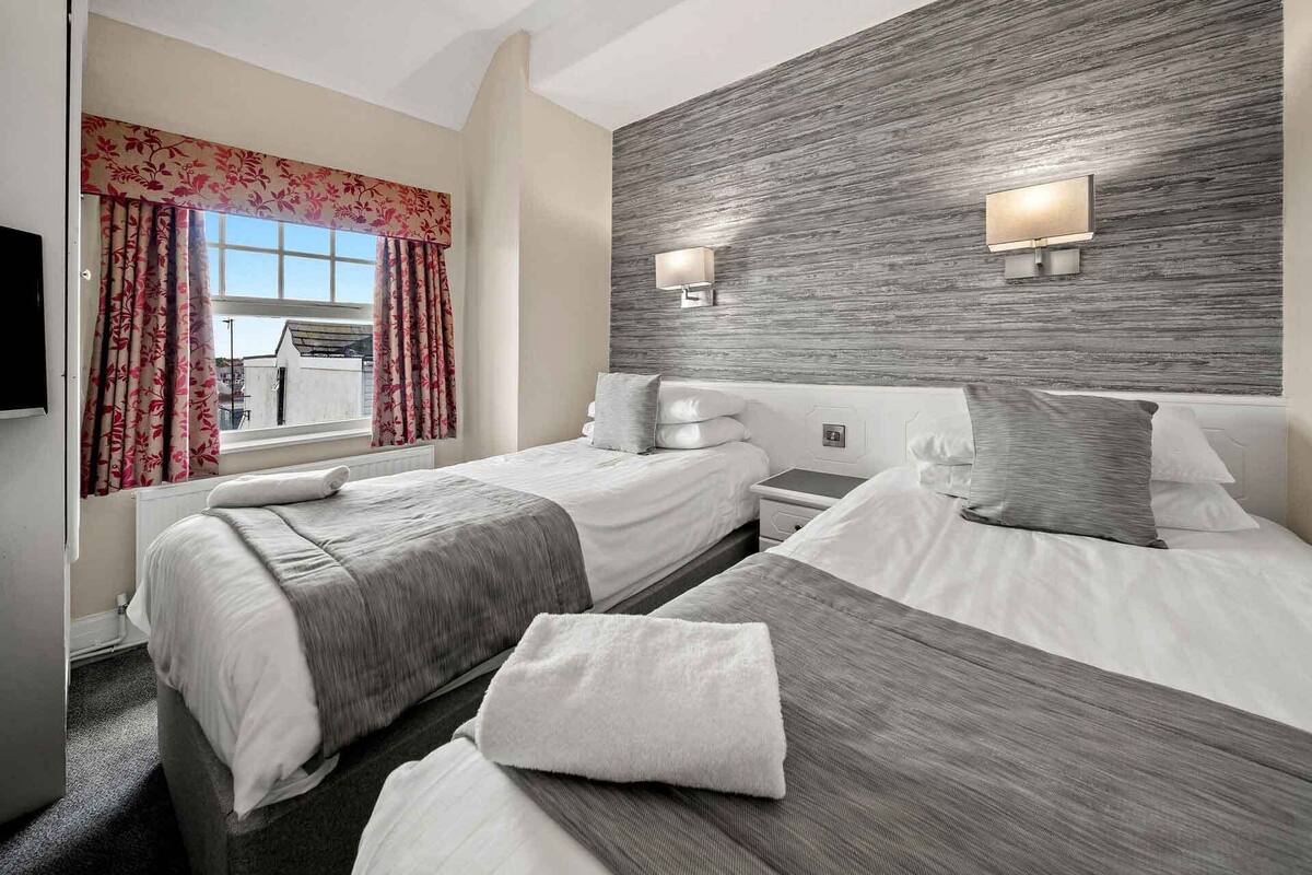 Habitación de hotel con dos camas, un ventanal con cortinas florales, una pared de detalles de madera e iluminación en la pared.