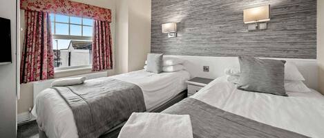 A hotel room with two beds, a large window with floral curtains, a wooden accent wall, and wall-mounted lighting.