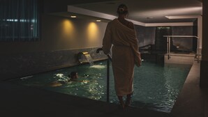 Una persona con bata de baño al lado de una piscina, con vista a un paisaje urbano a través de una ventana.