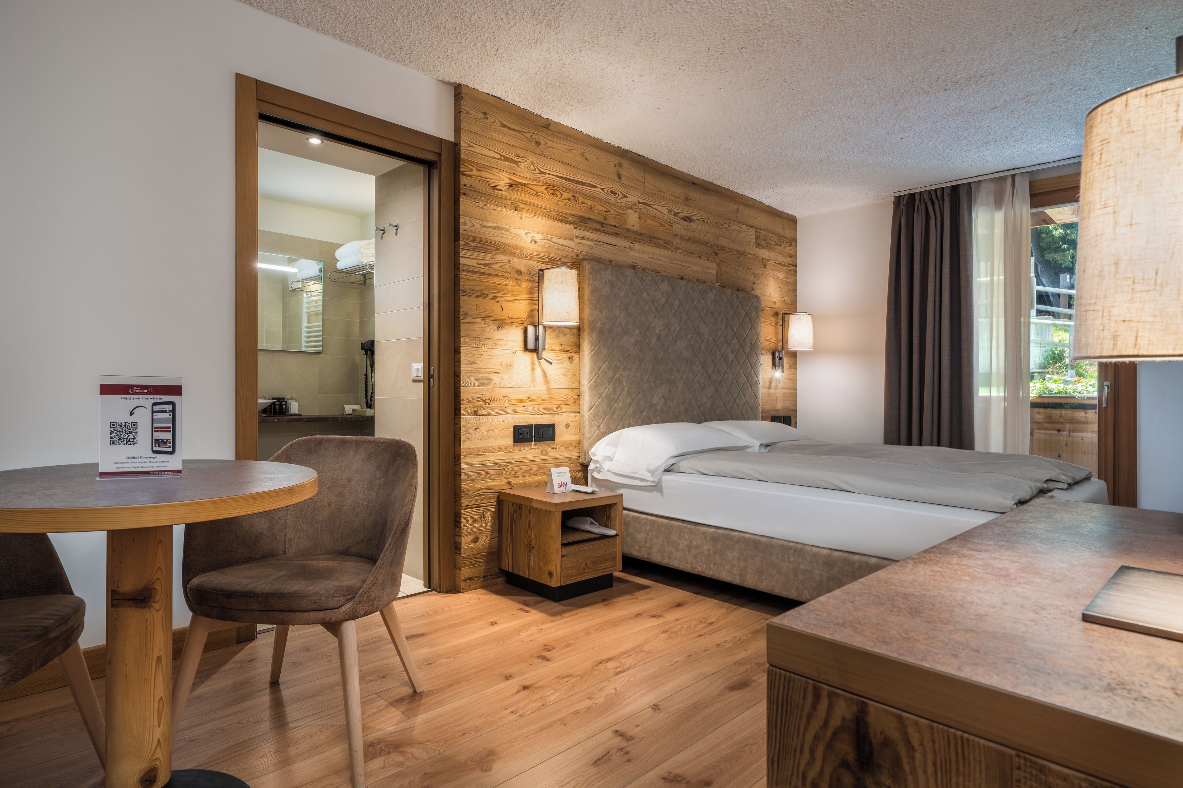 Habitación de hotel con cabecera de madera, una cama, una mesita de noche, una silla y una mesa redonda.