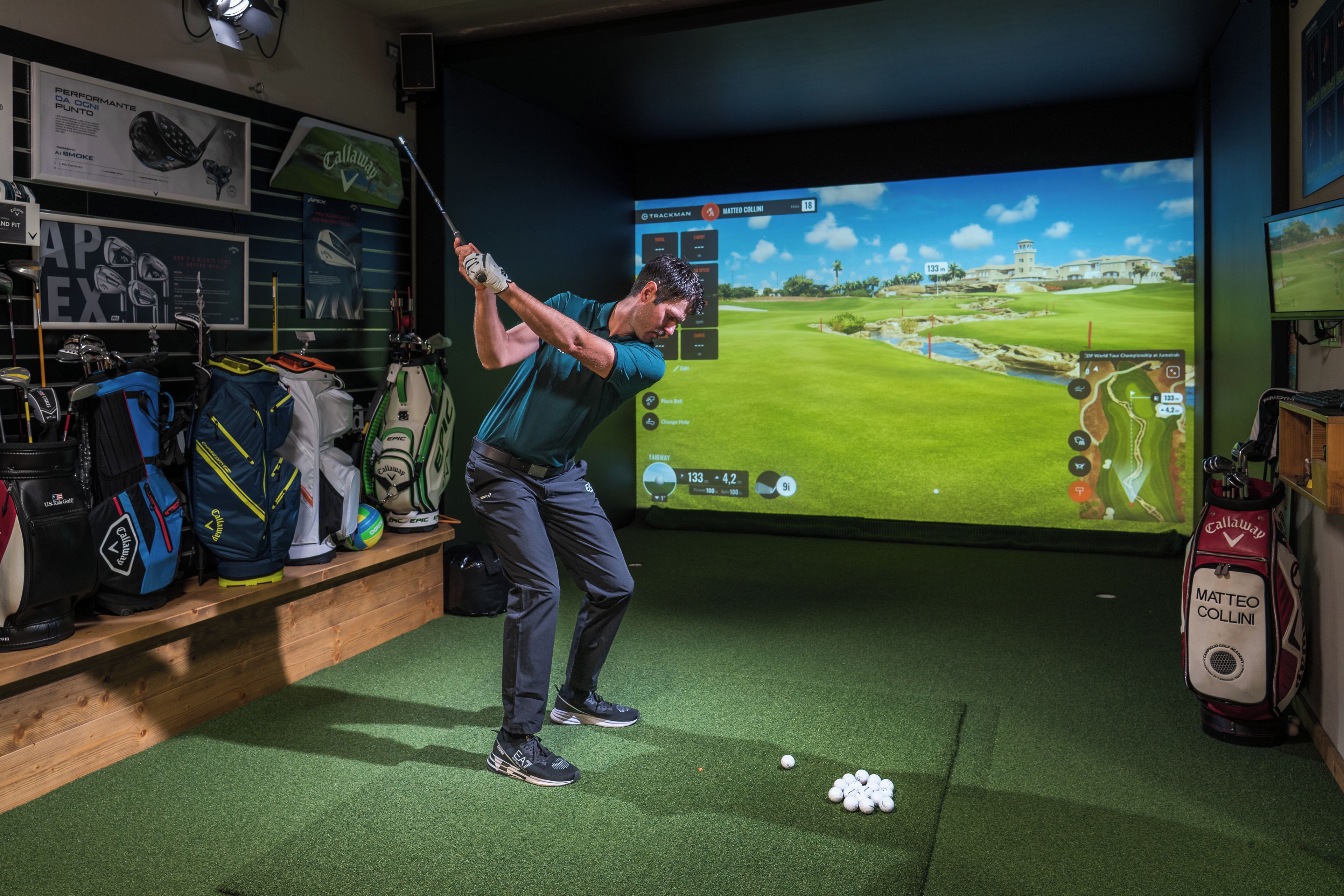 Configuración de simulación de golf con pantalla mostrando un campo de golf, un bolso de golf con la etiqueta 'Matteo Collini' y diverso equipo de golf en estanterías.