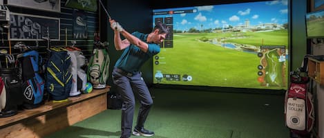 Configuración de simulación de golf con pantalla mostrando un campo de golf, un bolso de golf con la etiqueta 'Matteo Collini' y diverso equipo de golf en estanterías.