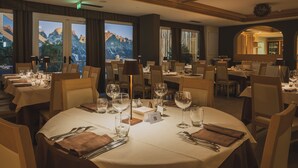 A restaurant with multiple tables set for dining, large windows with mountain views, and a warm interior lighting.