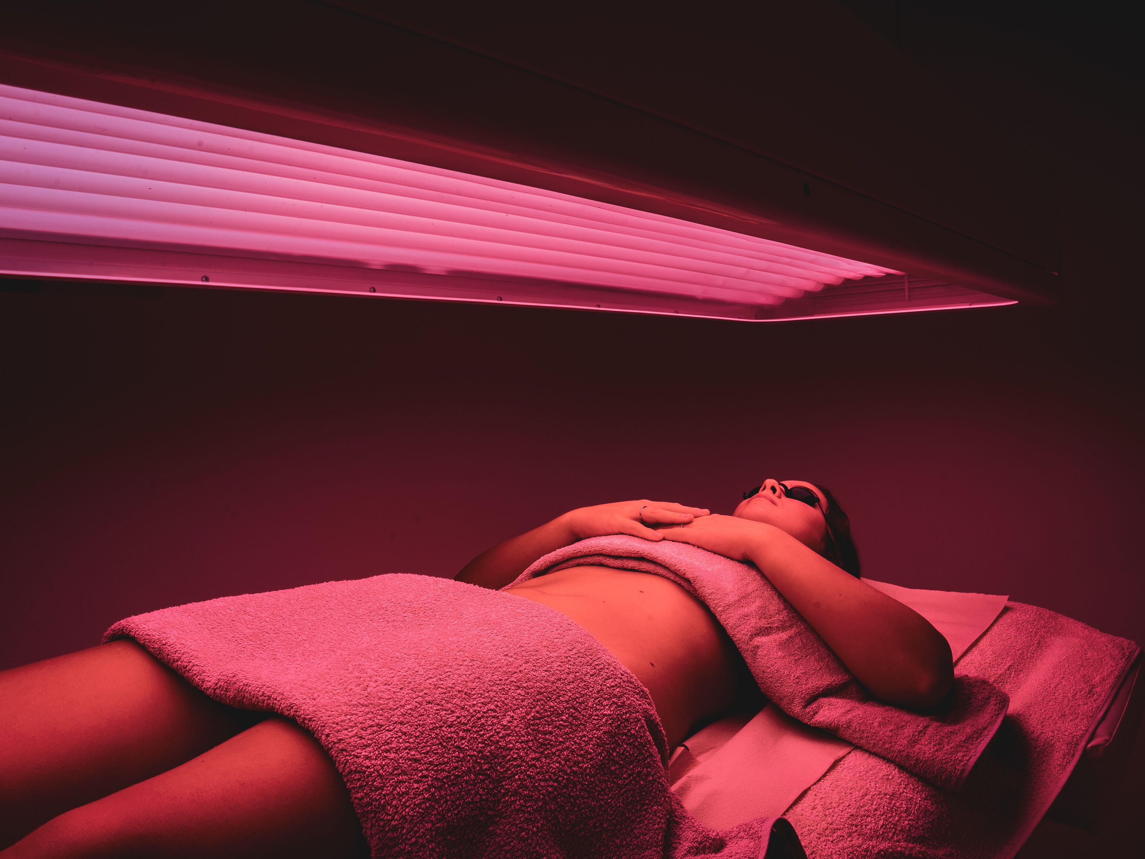 A person lying down with a towel covering their lower body, under a pink-lit ceiling.