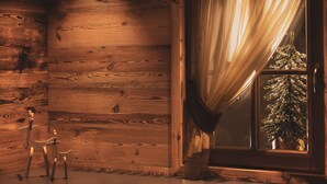 Interior de una cabaña de madera con ventana, cortina, un pequeño árbol de Navidad y una silla de madera.