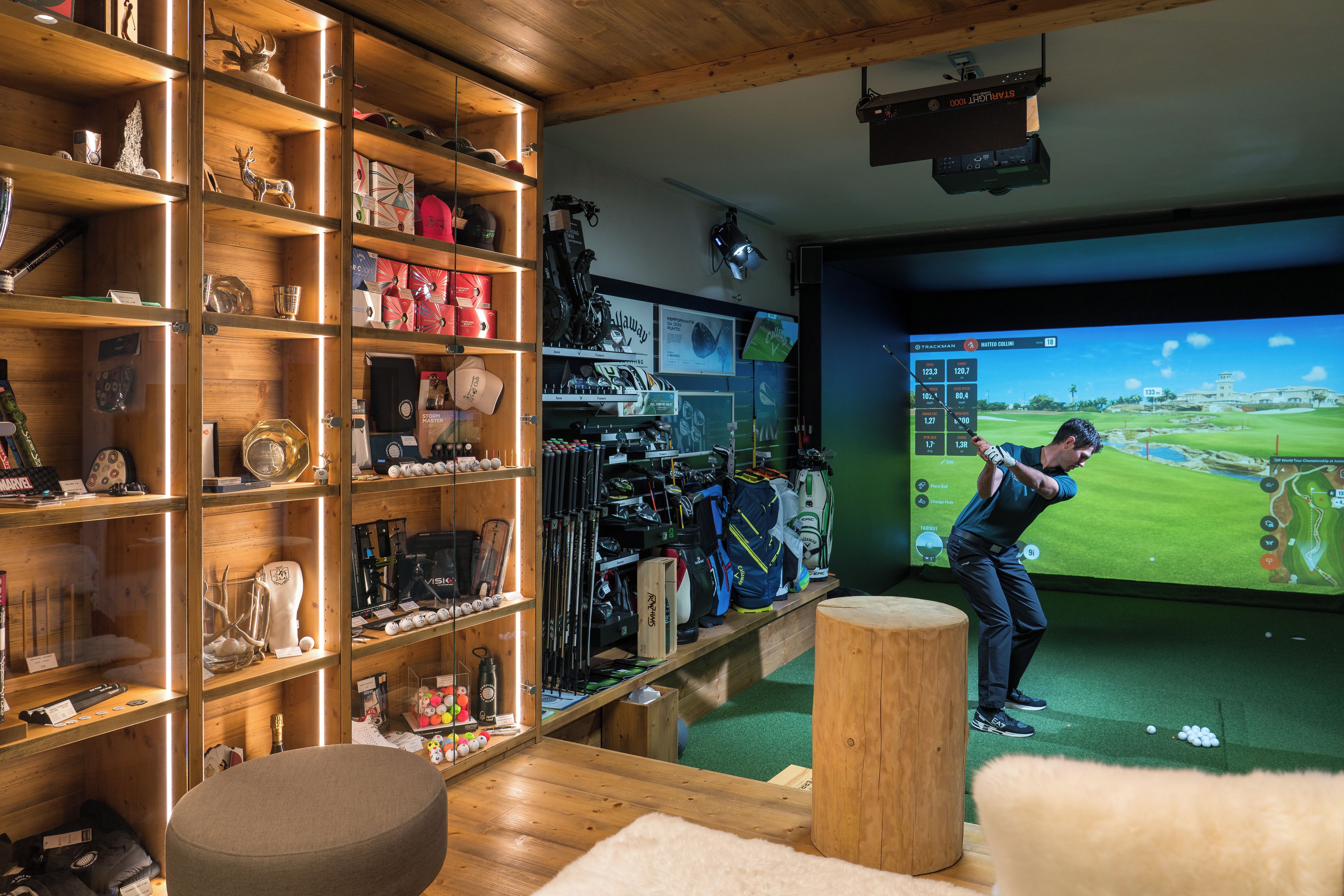 A room with a golf simulation setup, including a screen displaying a golf game, shelves with golf equipment, and a wooden floor.
